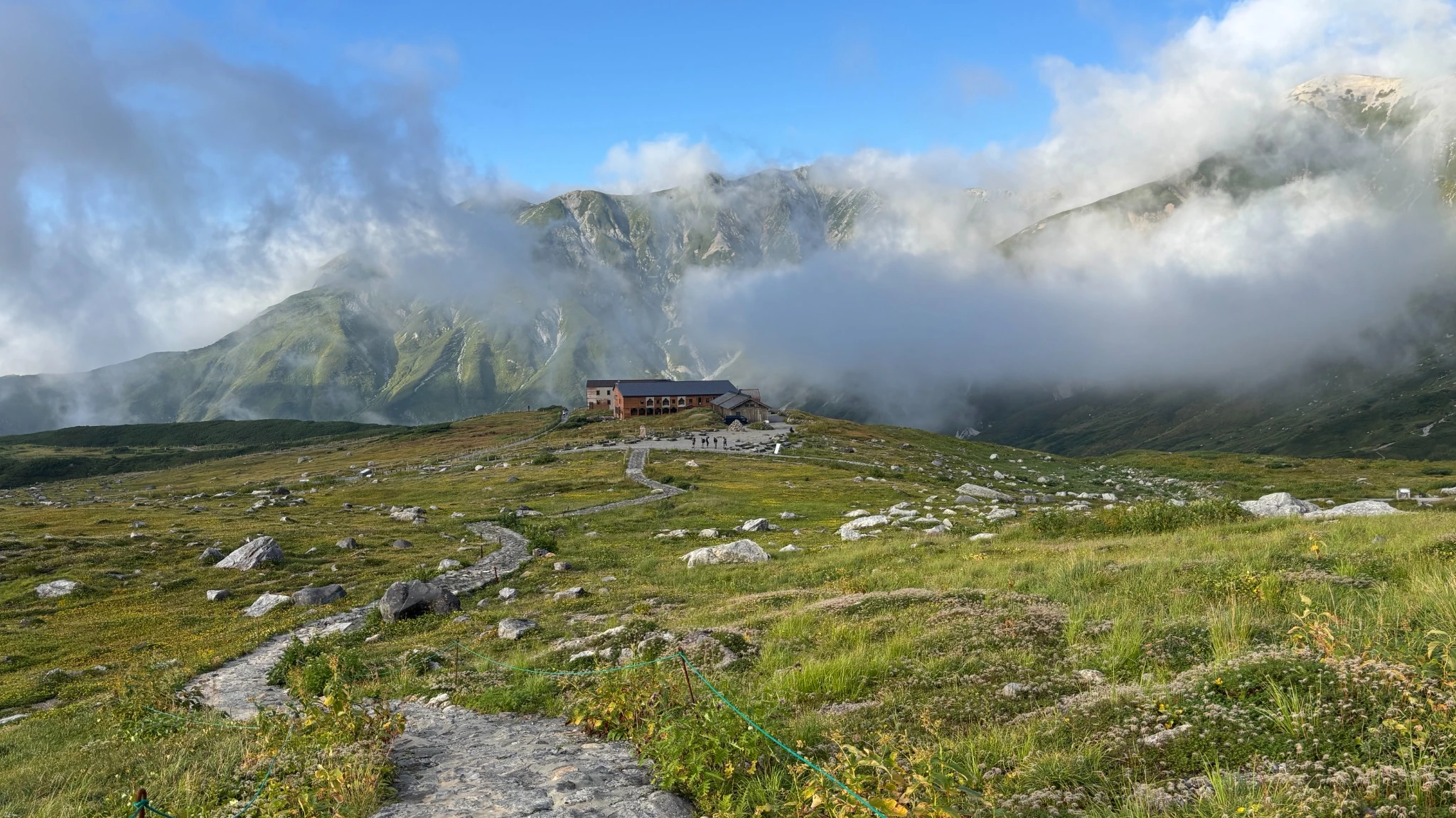 Stone path through the Murodo plateau toward a mountain lodge and cloud-wrapped peaks